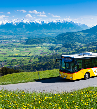 Ein Postauto auf einer Landstrasse mit grossen Wiesen und Bergen im Hintergrund